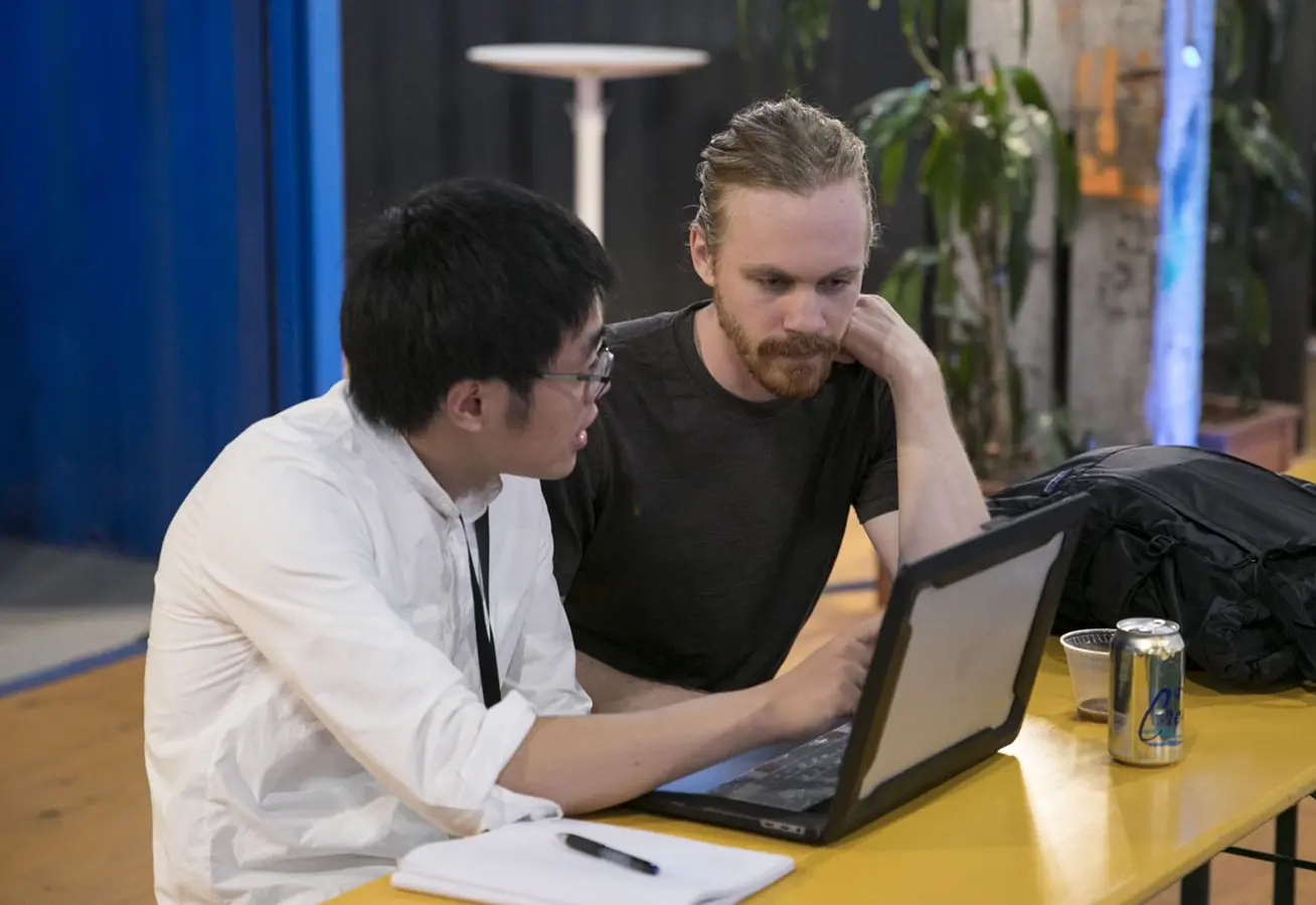 Two people working closely on a laptop at a table, discussing code with a notebook and drink nearby.